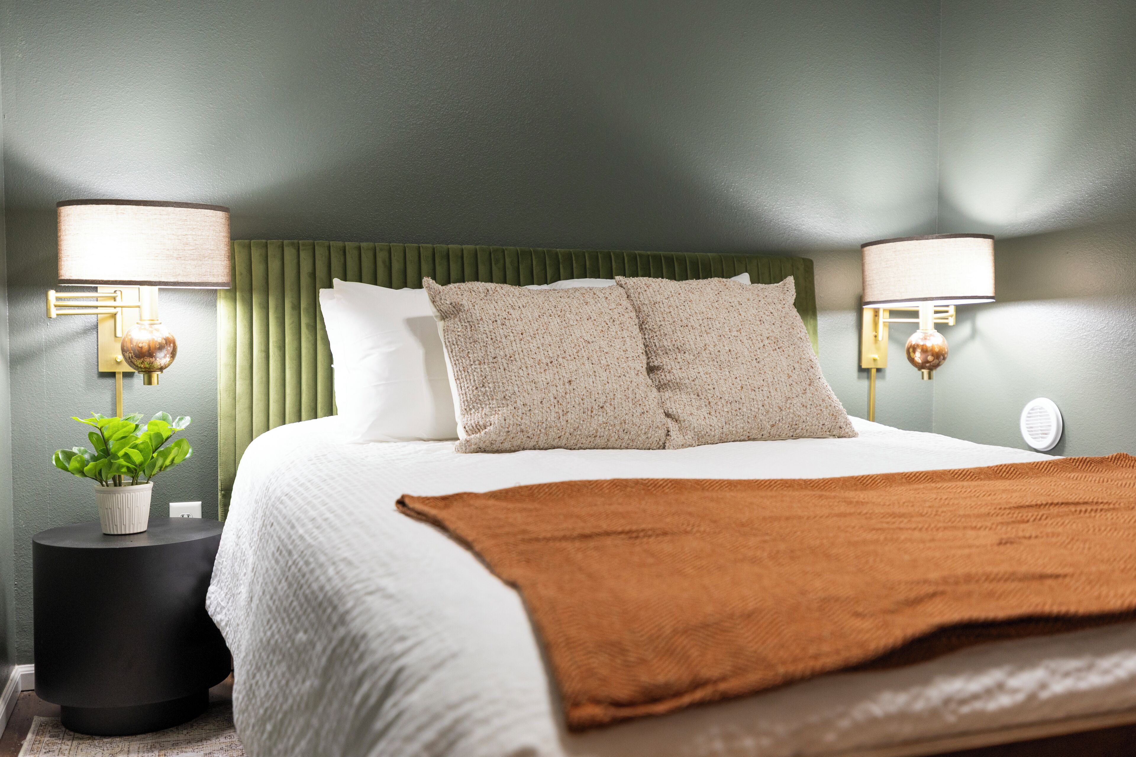 A neatly made bed with a green headboard, two bedside lamps, and a potted plant on a side table.