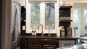 A kitchen with dark wood cabinets, a stainless steel refrigerator, a double sink with a gold faucet, and a window with a view of trees. - The Peak: Hot tub + Mt. Views (Packwood)