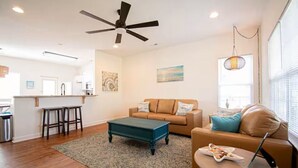 A spacious living room with a dark blue coffee table, a beige sofa, and a tan couch. There is a ceiling fan, a kitchen in the background, and a large window with blinds.