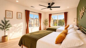Habitación de hotel con dos camas, ventilador de techo, ventanas amplias con cortinas, una planta en maceta y cuadros en las paredes.