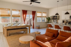 A modern living room with a wooden dining table set for a meal, a sofa, and a kitchen area with a TV and bar.
