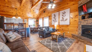 A cozy cabin interior with a wooden ceiling, a stone fireplace, a dining area, and a living room furnished with a sofa and a coffee table.
