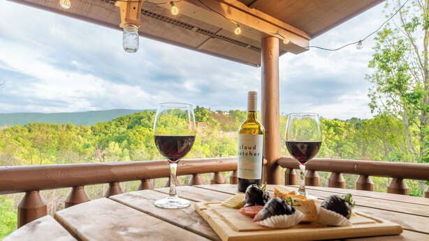 A wooden table with a bottle of wine, two glasses of red wine, and a board with cheese and crackers on a balcony with a scenic view.