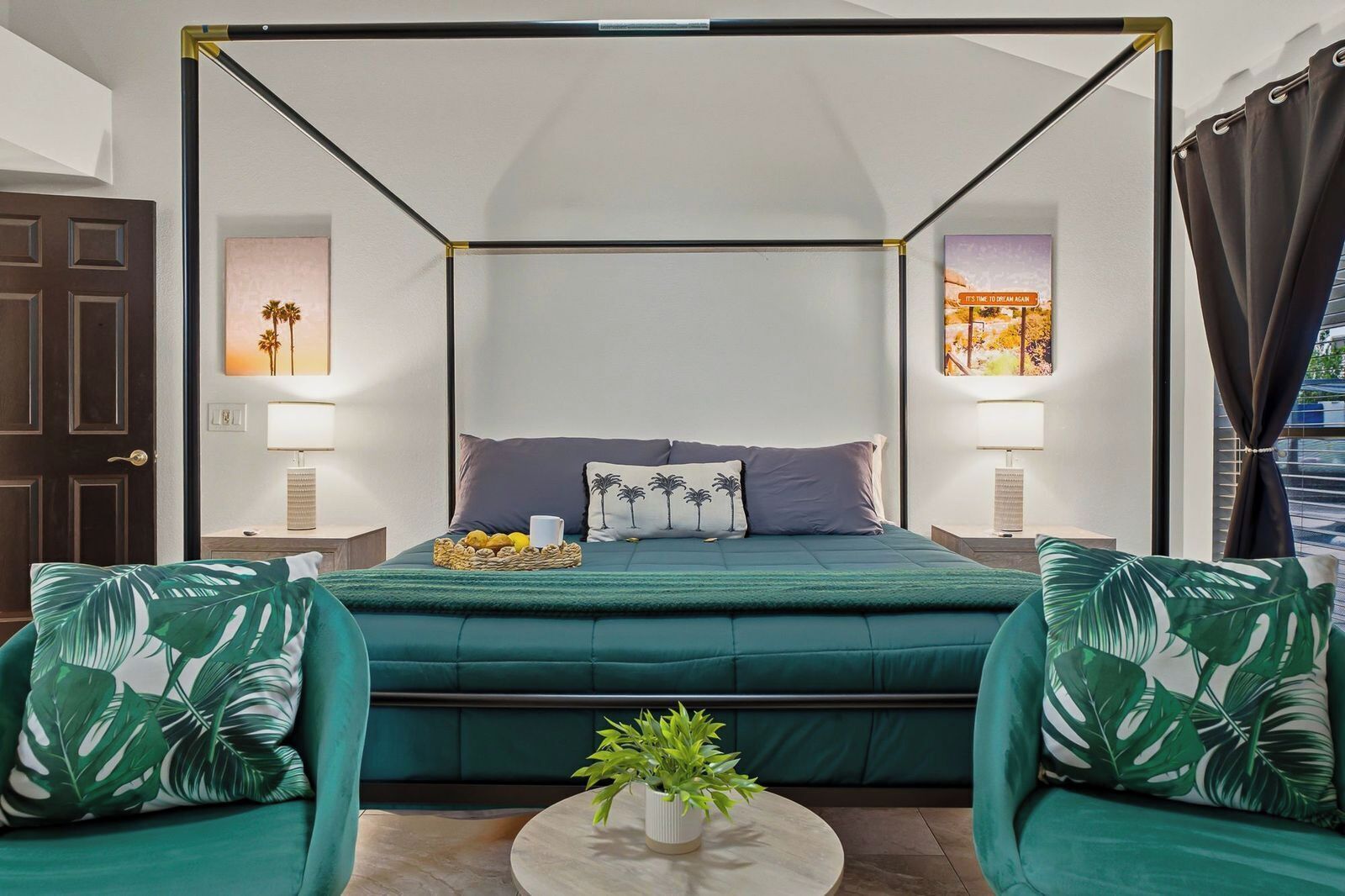 A four-poster bed with a green upholstered headboard, two nightstands with lamps, a small table with a plant, and a door with a dark frame.
