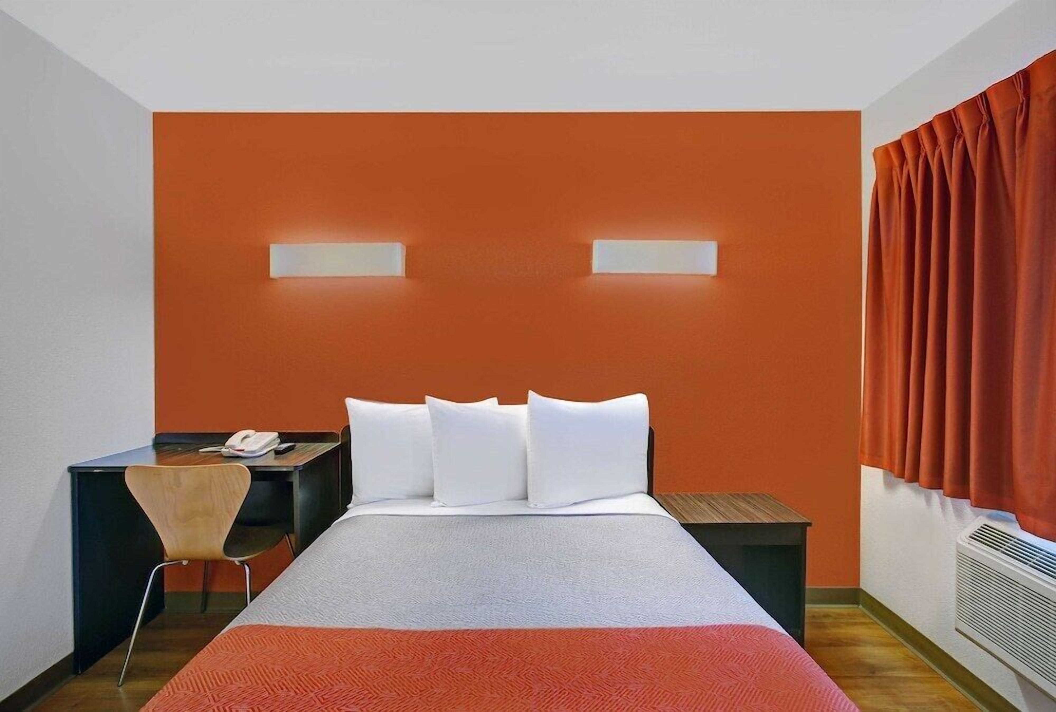 Une chambre d’hôtel avec un mur d’accent orange, un lit avec une literie blanche, un bureau avec une chaise et une fenêtre avec des rideaux orange.