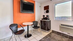 Ett litet rum med ett vitt bord, två stolar, en tv som hänger på en orange vägg, en kaffemaskin och ett fönster med utsikt.