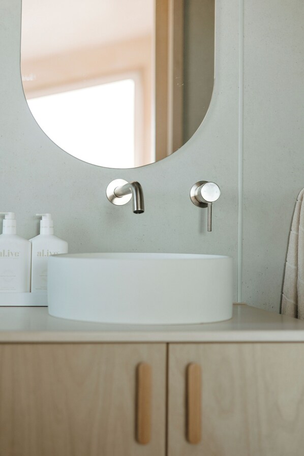 A bathroom with a round mirror, a white sink, and wooden cabinets. - Freya - Seaside Tiny House With Sweeping Ocean Views (North Cape)
