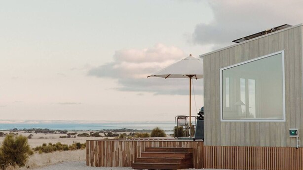 Ein modernes Gebäude mit großem Fenster, einer Holzterrasse und Blick auf Strand und Meer.