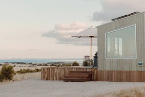 A modern building with a large window, wooden deck, and a view of the beach and ocean. - Freya - Seaside Tiny House With Sweeping Ocean Views (North Cape)