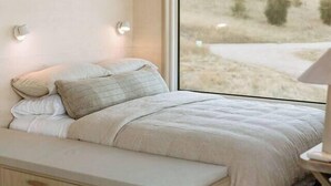 Ein Schlafzimmer mit großem Fenster, einem Bett mit Kissen, einem Nachttisch und einer Bank.