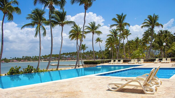 A pool with lounge chairs, palm trees, and a beach in the background.