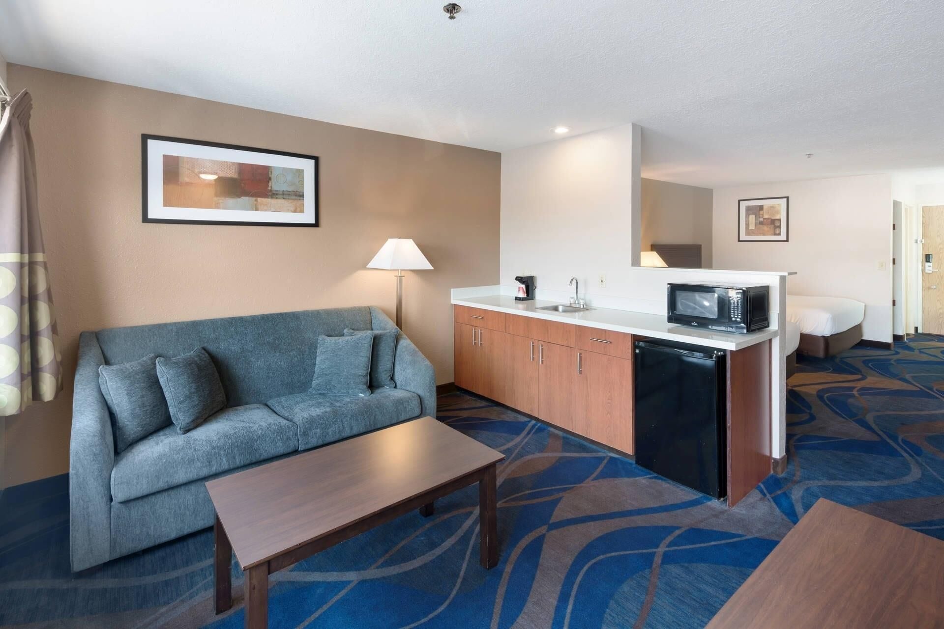 A hotel room with a sofa, a small table, a kitchenette with a microwave, and a bed in the background.
