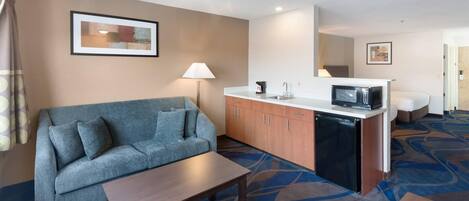 Habitación de hotel con sofá, una mesita, kitchenette con microondas y una cama al fondo.