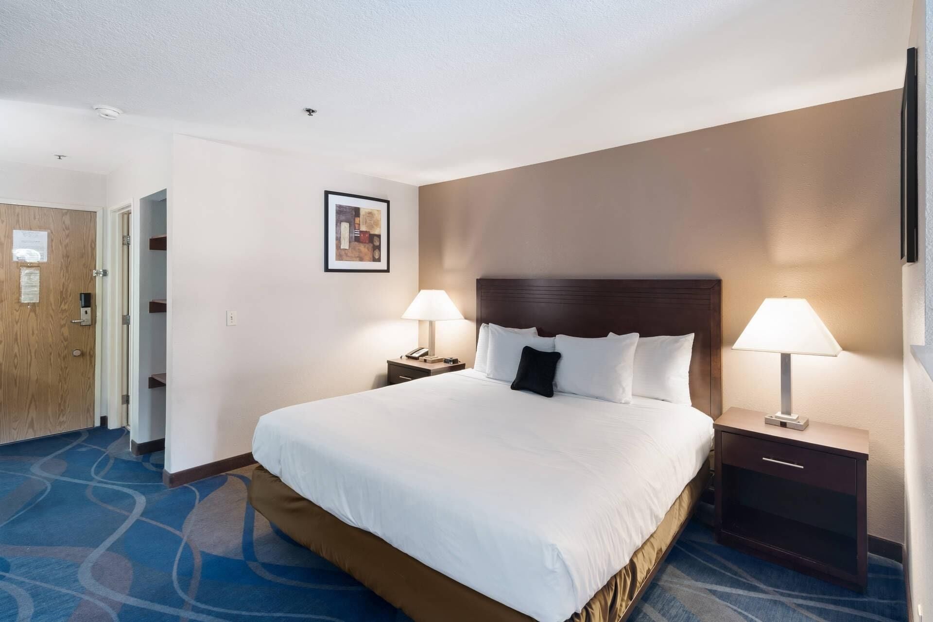 A hotel room with a large bed, two bedside tables with lamps, a wooden door, and a framed picture on the wall.