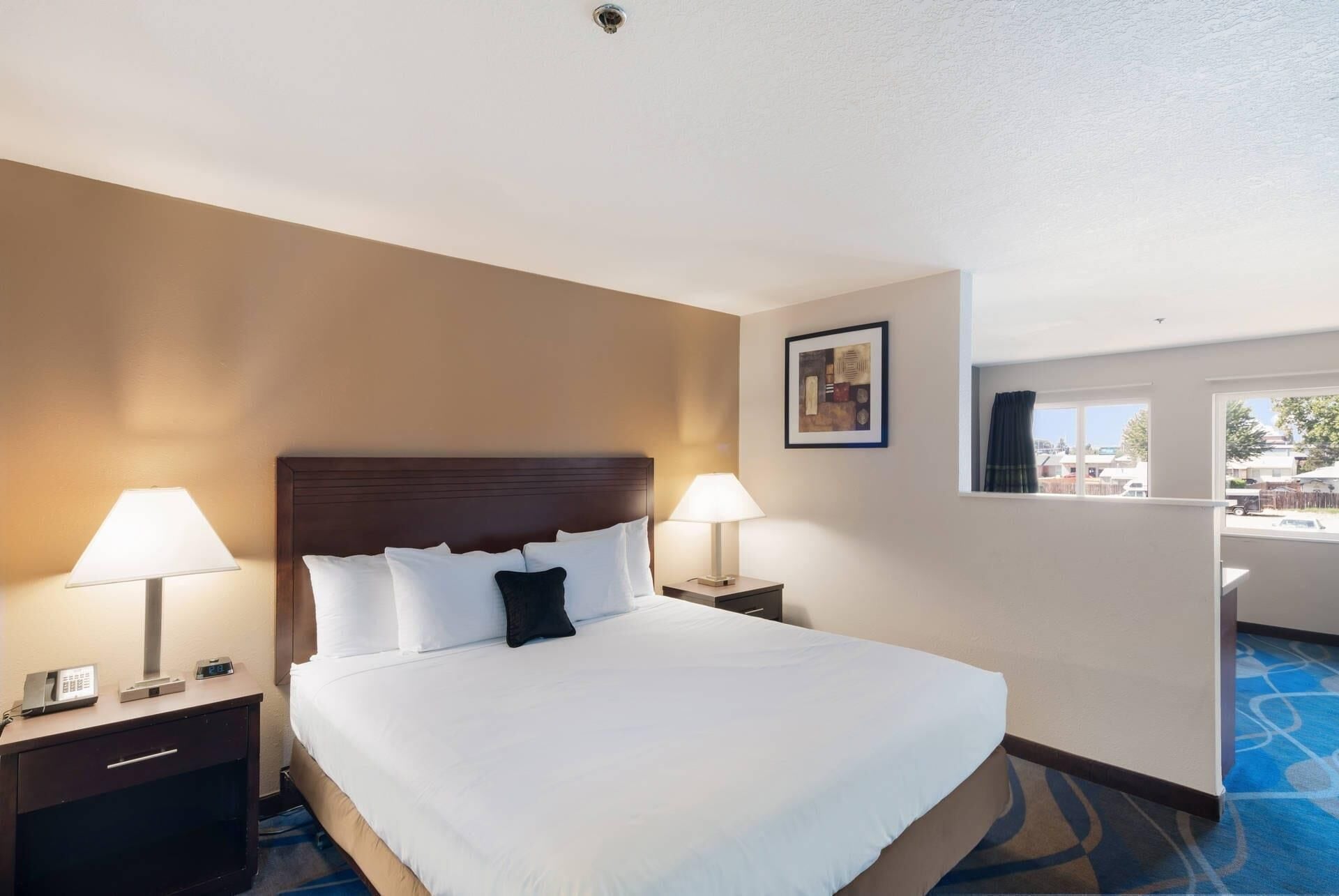 A hotel room with a large bed, two bedside tables with lamps, a painting on the wall, and a view through a window.