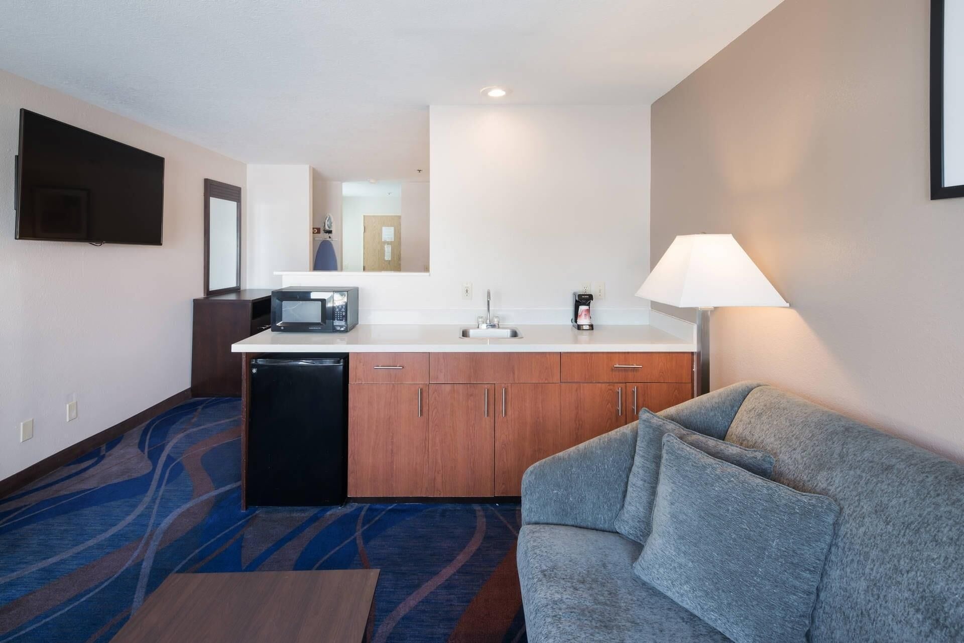 A compact hotel room with a kitchenette, a flat-screen TV, a sofa, and a lamp.