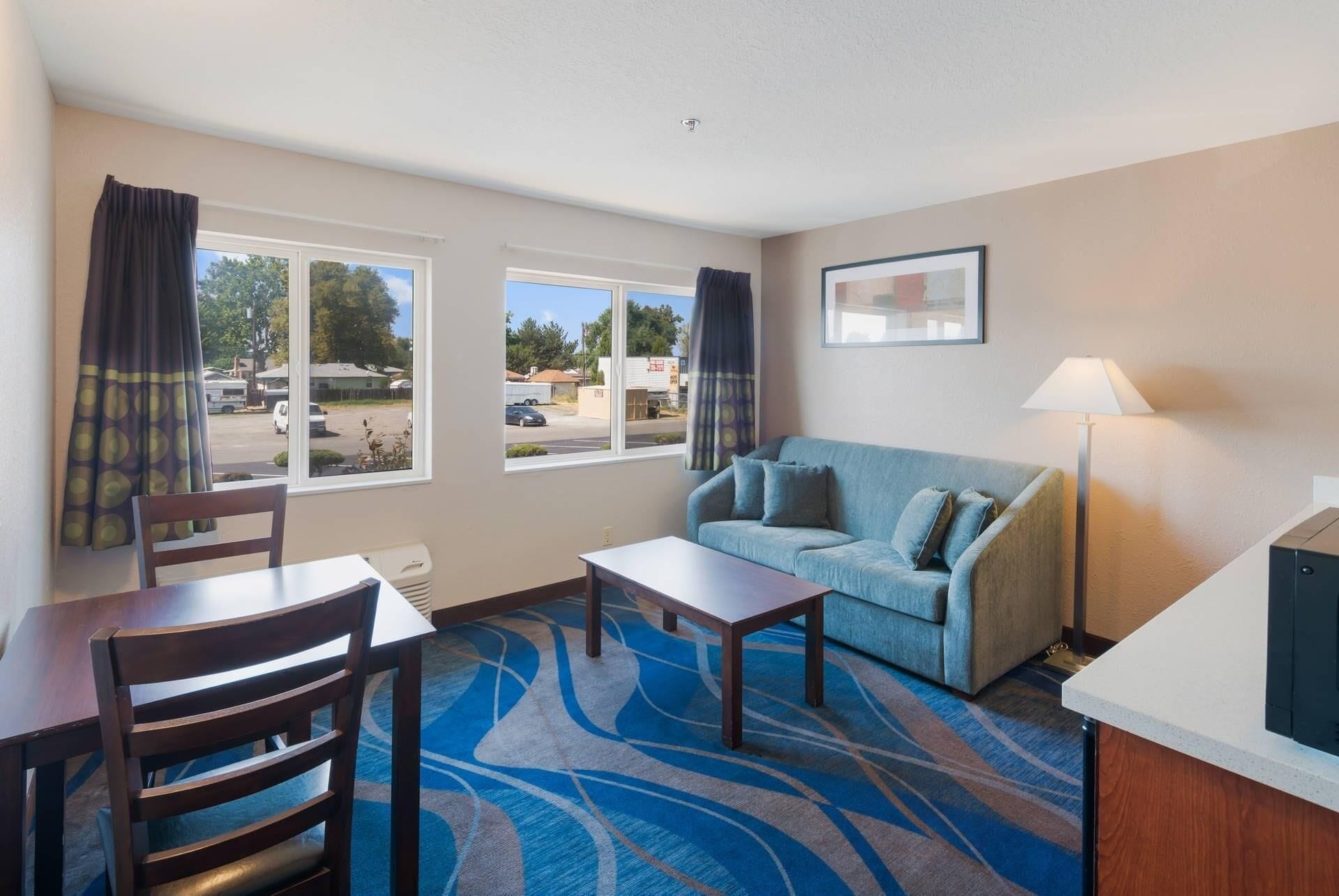 A hotel room with a sofa, a dining table with chairs, a small table, and a lamp. There are two windows with curtains, and a framed picture on the wall.