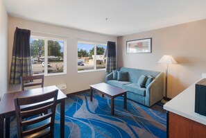 Habitación de hotel con sofá, mesa de comedor con sillas, una mesita y una lámpara. Hay dos ventanas con cortinas y un cuadro en la pared.