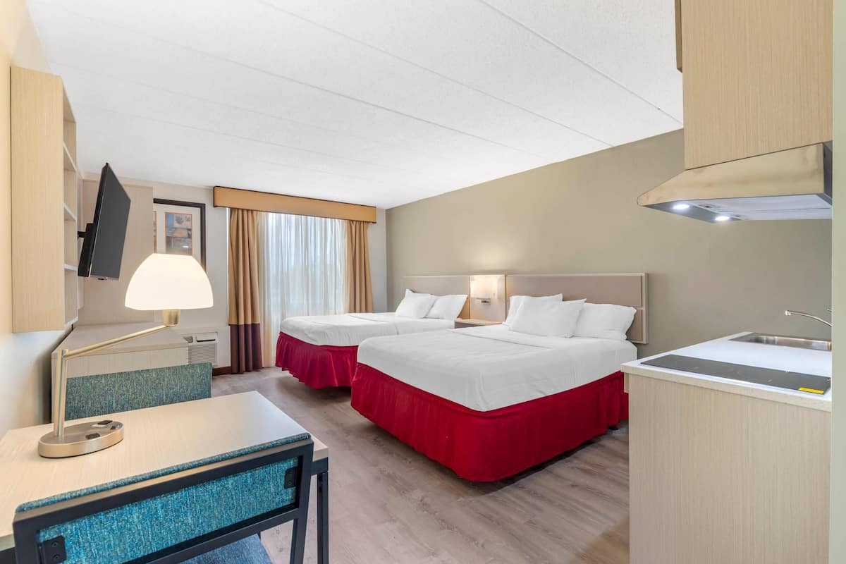 A hotel room with two beds, a desk, a chair, a lamp, a TV, a window with curtains, and a kitchenette.