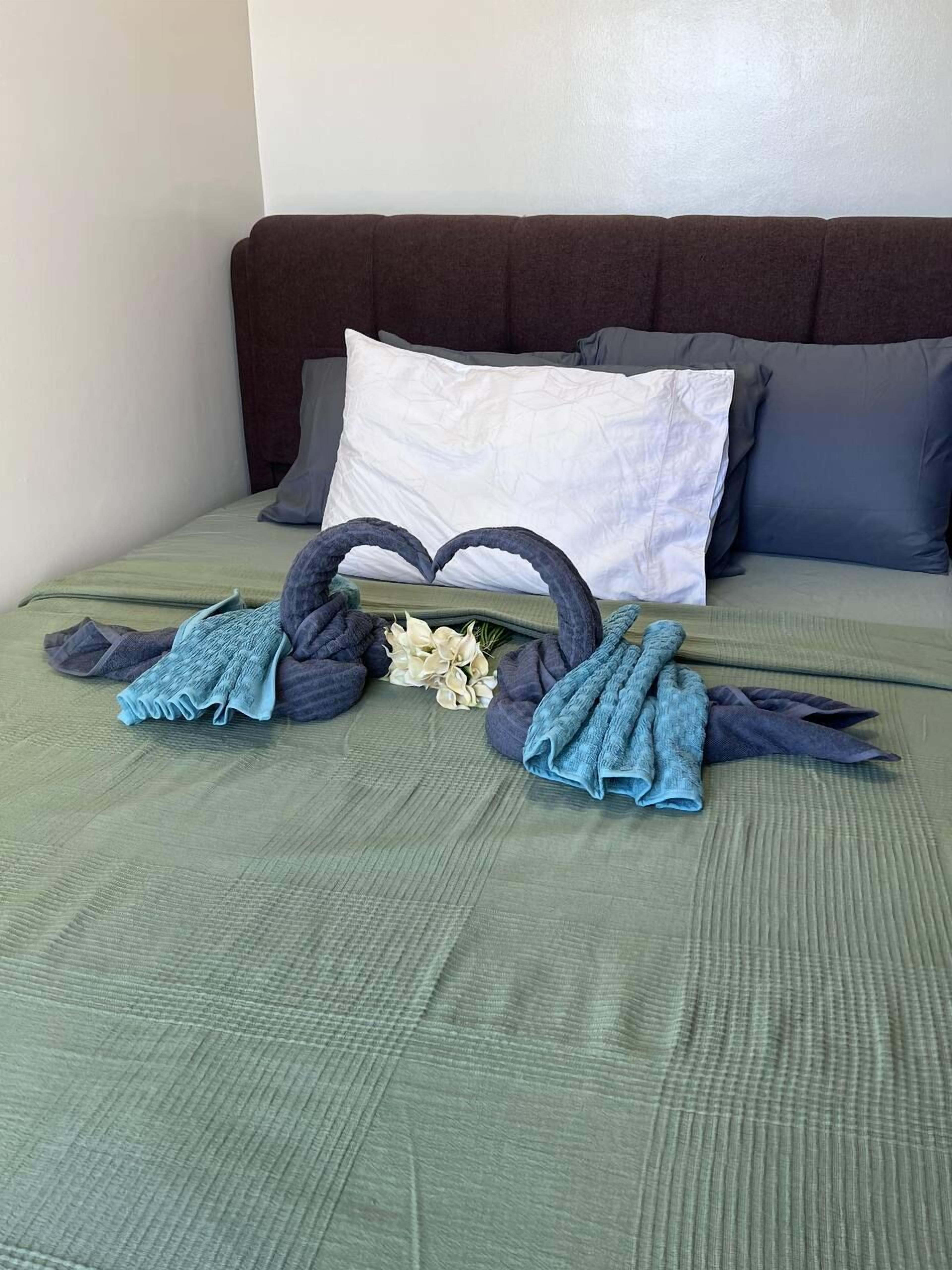 A neatly made bed with a green bedspread, two heart-shaped towels, and a small bouquet of flowers.