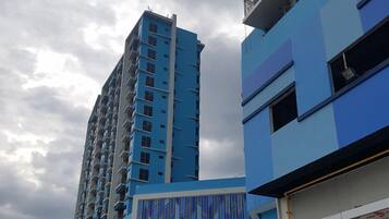 A modern building with a blue facade, multiple balconies, and a parking area with a white van and a red car.