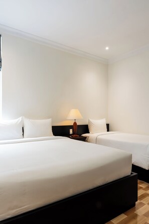 Habitación de hotel con dos camas, una mesita de noche con lámpara y suelo de madera.
