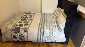 Una habitación pequeña con cama individual, piso de madera, cabecera negra y ropa de cama estampada.