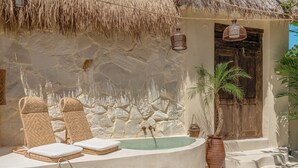 A stone wall with a thatched roof, a wooden door, a bathtub, and two wicker chairs with cushions.