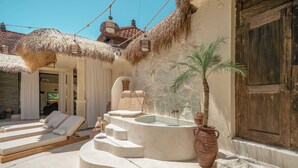 A sunlit outdoor lounge area with a stone bench, a hot tub, and a palm tree.