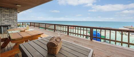 A wooden deck with picnic tables and a wicker basket, overlooking a beach with a clear view of the ocean and distant buildings.