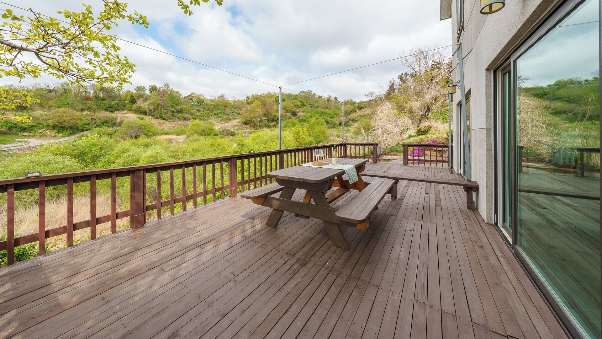 Une terrasse en bois, dotée de tables de pique-nique et d’une rambarde en verre, qui offre une vue sur un paysage naturel.