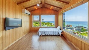 A bedroom with a bed, a TV, and large windows with a view of the ocean and greenery. - Gyeongju Happy Dream Ocean View Pension (Gyeongju)