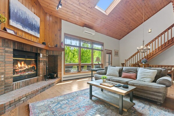 A cozy living room with a fireplace, a large window, a staircase, and a modern sofa.