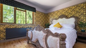 A bedroom with a large bed, ornate wooden headboard, patterned wallpaper, and a window with a view of trees. - Sampford Hideaway Retreat (Little Sampford)