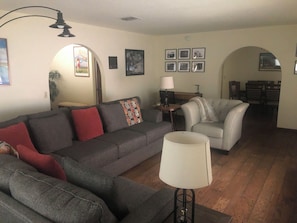 Una sala de estar con un sofá gris, un sillón blanco, una lámpara y un comedor al fondo.