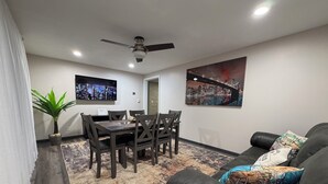 A modern living room with a dining area, a ceiling fan, a flat-screen TV, and a cityscape painting. - Charming 3 Bedroom 2.5 bathroom finished basement, Huge yard and Charcoal grill (Baraboo)