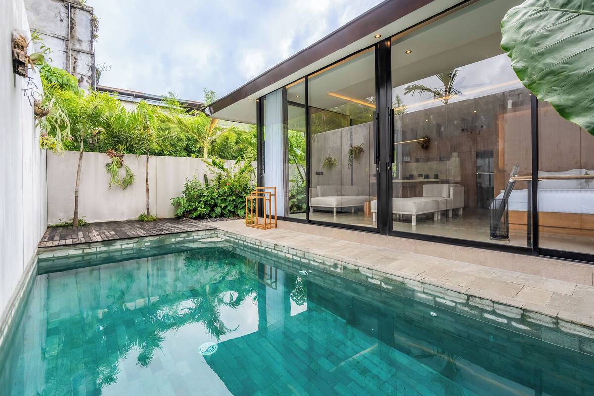 Ein moderner Poolbereich im Freien mit einer verglasten Terrasse, einer Sitzecke mit Sofa und Sesseln sowie üppiger Bepflanzung.