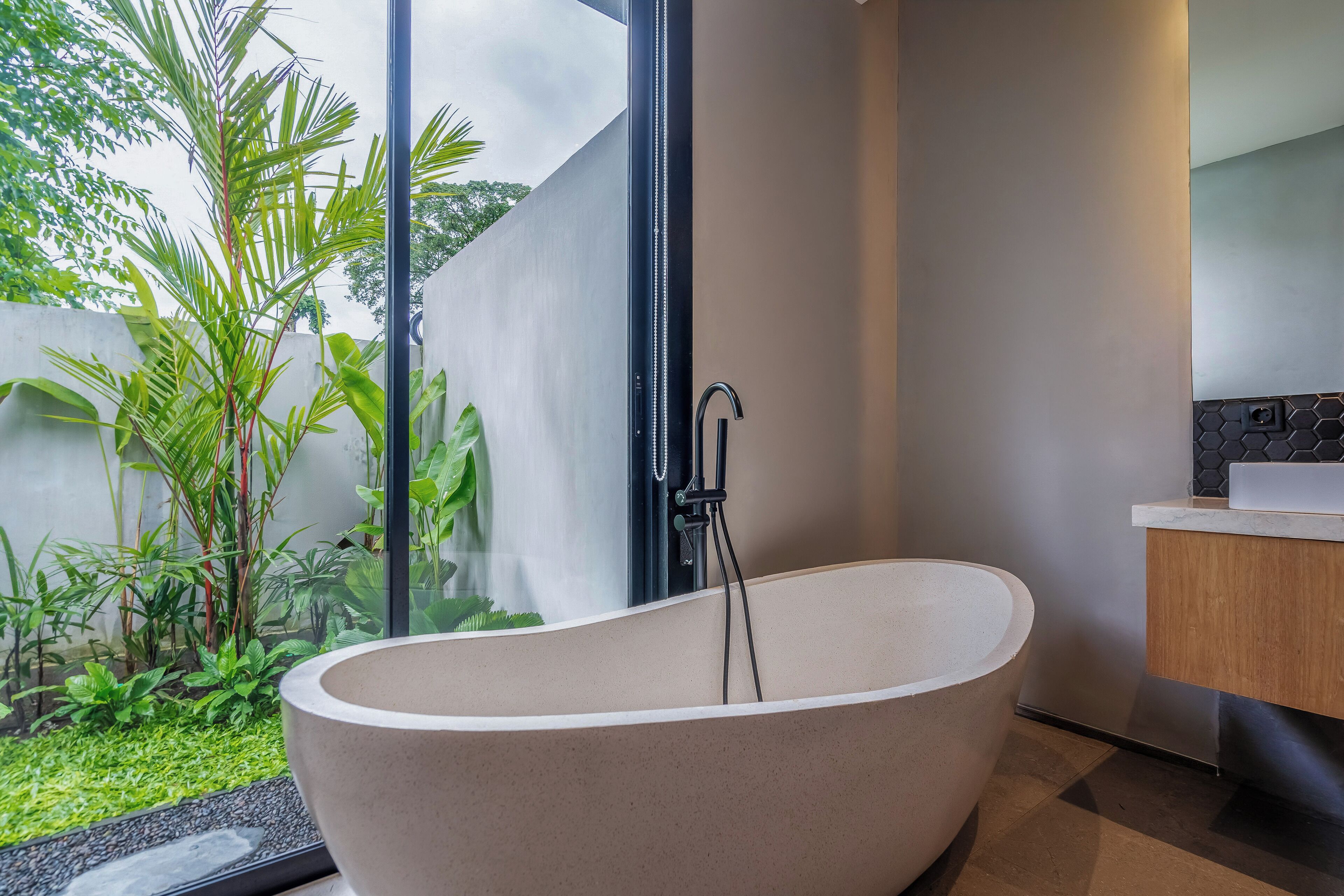 A modern bathroom with a freestanding bathtub, a wooden vanity, and a view of a garden.