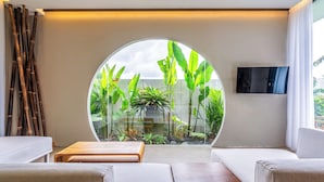 A modern living room with a circular glass window, a flat-screen TV, and a seating area with sofas and a coffee table. - Titi House Villa By Betterplace (Ubud)