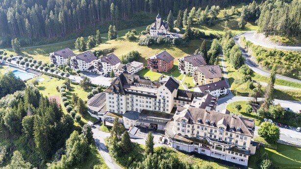 Vista aérea de un complejo hotelero con varias construcciones, una piscina y un área boscosa al fondo.