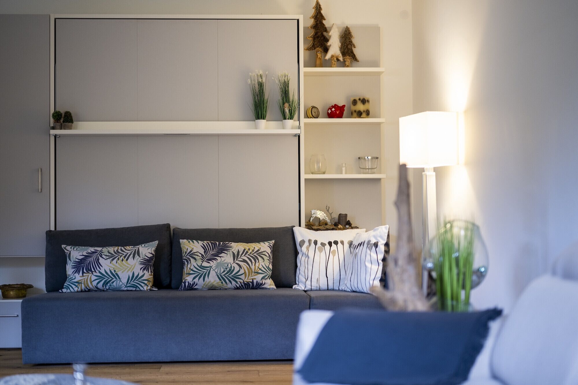 Una sala moderna con un sofá gris, cojines decorativos, un mueble blanco y una estantería con objetos pequeños y una lámpara.