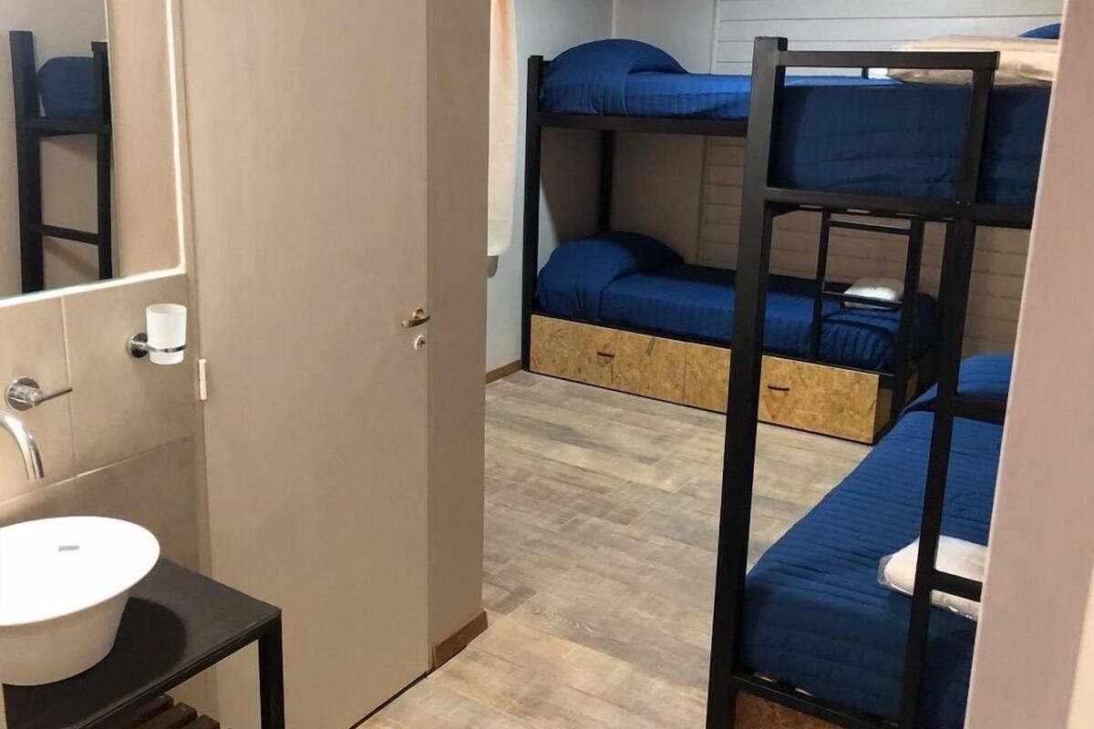 Une chambre de dortoir avec des lits superposés, un plancher en bois, une petite salle de bain avec un lavabo et un miroir, et une porte donnant sur une autre pièce.