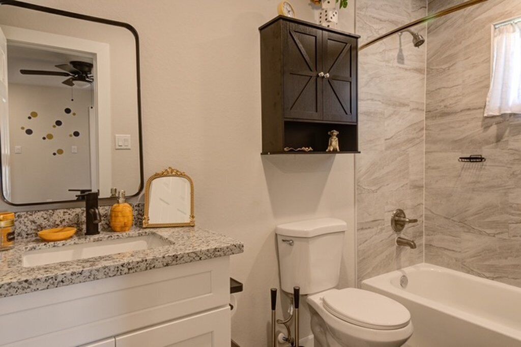 A bathroom with a marble wall, a white toilet, a bathtub, a sink with a granite countertop, a dark wooden cabinet, and a mirror with a gold frame.