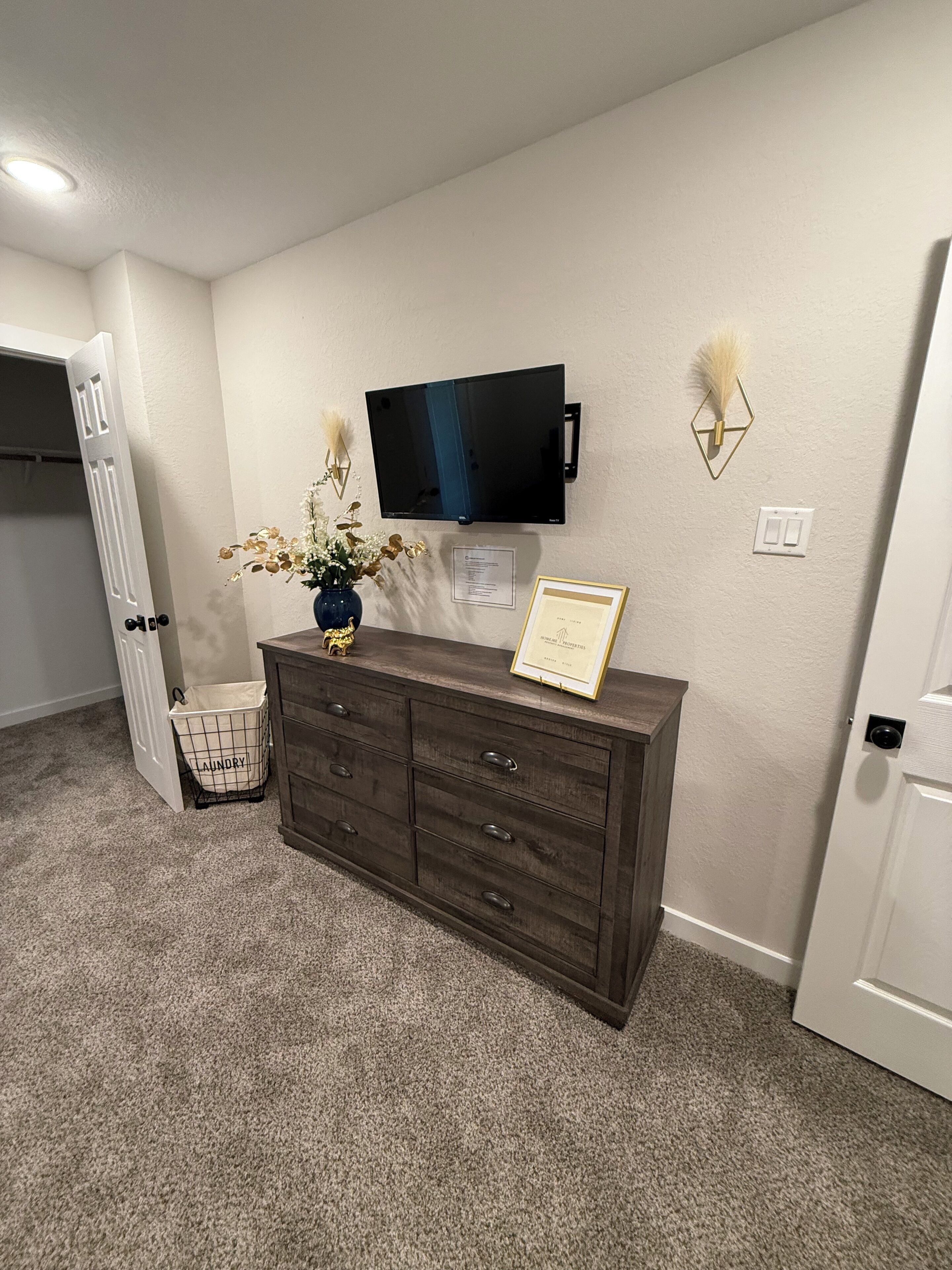 A room with a dark wooden dresser, a flat-screen TV mounted on the wall, a vase with flowers, and a laundry basket labeled "laundry".