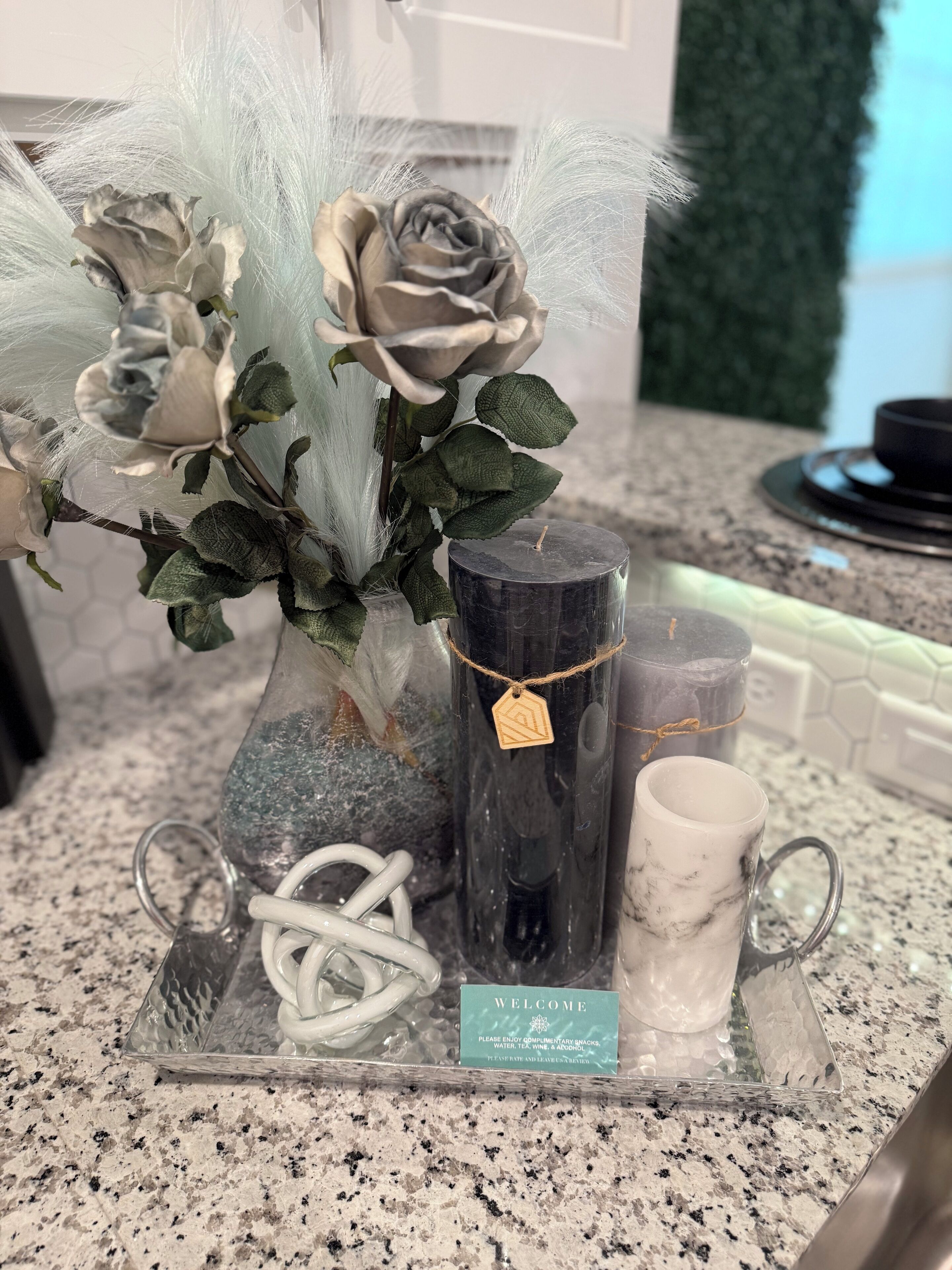 A clear glass tray with a decorative arrangement including a vase with artificial flowers, a candle, a mug, and a welcome sign.