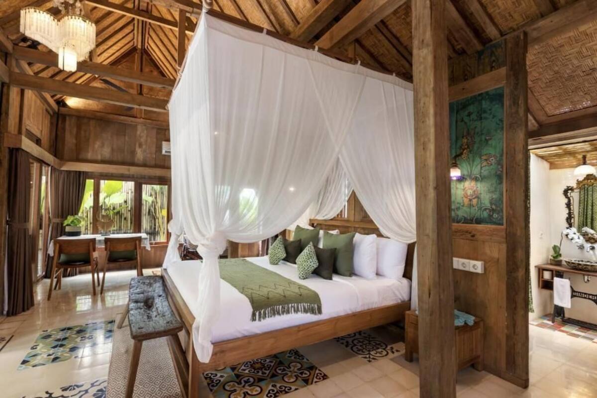 A spacious room with a large bed, wooden ceiling beams, and a view of greenery outside.