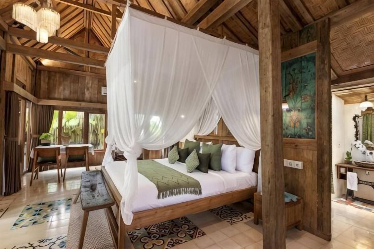 A four-poster bed with a canopy, a wooden floor, a dining area with a table and chairs, and a painting on the wall.