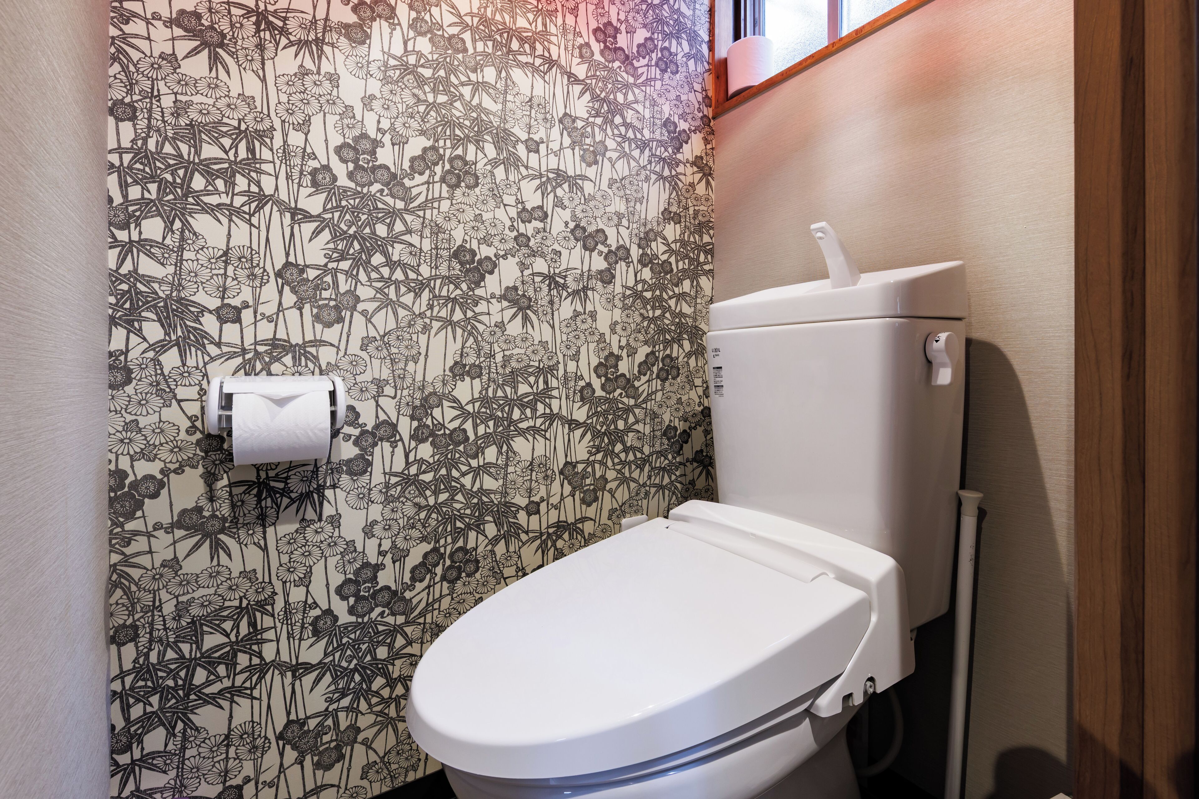 Des toilettes avec un abattant et une cuvette blancs, un porte-rouleau de papier toilette fixé au mur, et un papier peint à motifs floraux et de bambou.
