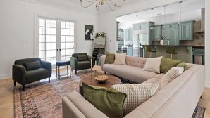 Ein geräumiges Wohnzimmer mit einem großen, beigefarbenen Ecksofa, zwei Sesseln, einem Couchtisch und einem gemusterten Teppich. Durch einen offenen Durchgang ist eine Küche mit türkisfarbenen Schränken zu sehen.