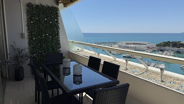 Un balcon avec une table et des chaises, donnant sur une marina où sont amarrés des bateaux, avec un ciel bleu dégagé.